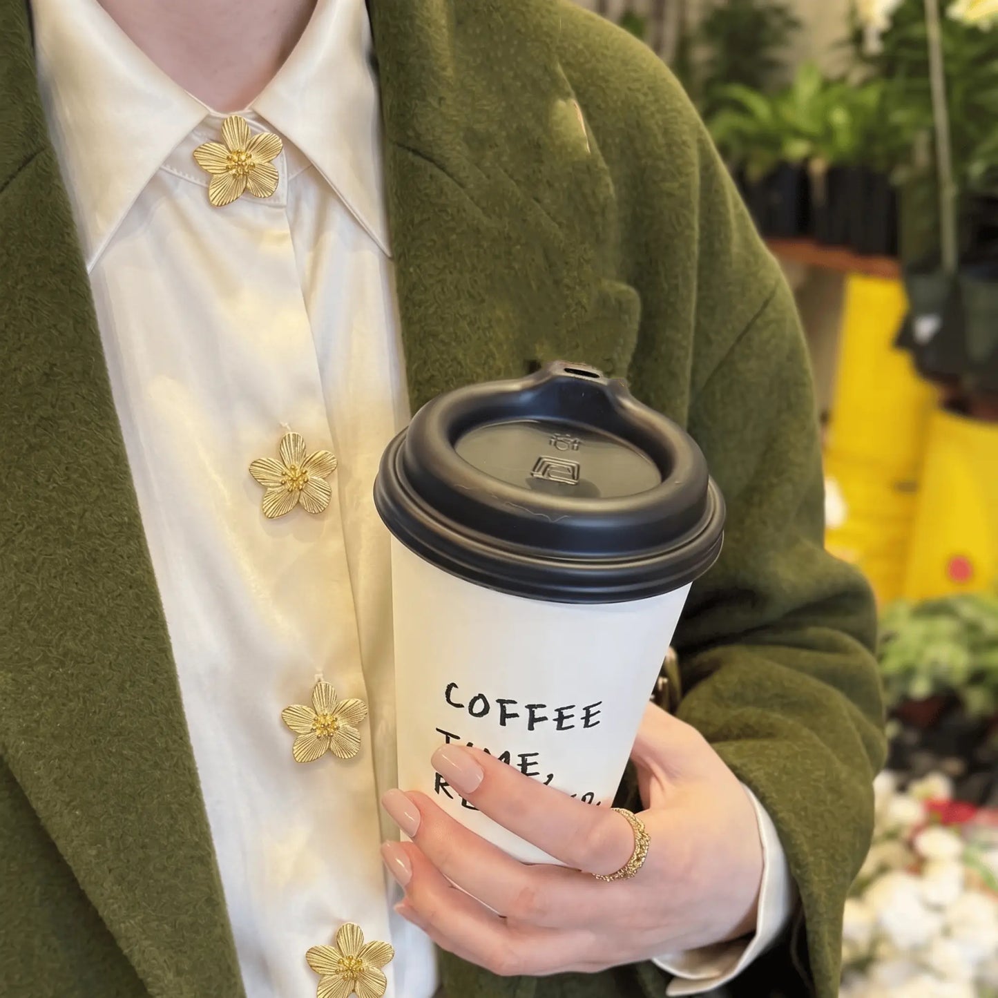 A chic everyday lifestyle shot of a woman on a coffee run, wearing an olive green coat over a white silk shirt. The shirt's front placket and top collar button are elegantly styled with realistically scaled textured gold floral button covers. A creative, light luxury faux cufflink alternative for sophisticated daily fashion.