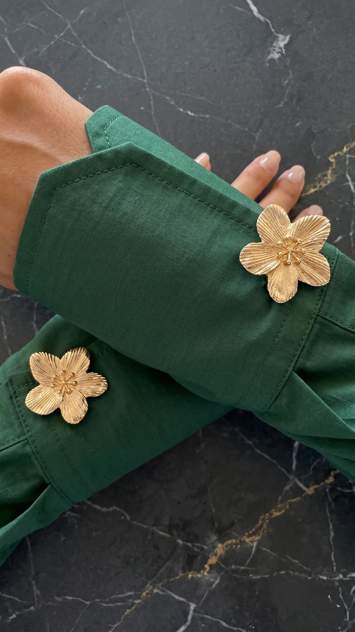An elegant top-down shot of crossed arms in a rich emerald green shirt, beautifully adorned with two textured gold floral button covers. The dark marble background emphasizes the luxurious brushed metal finish and sophisticated color contrast for modern power dressing.
