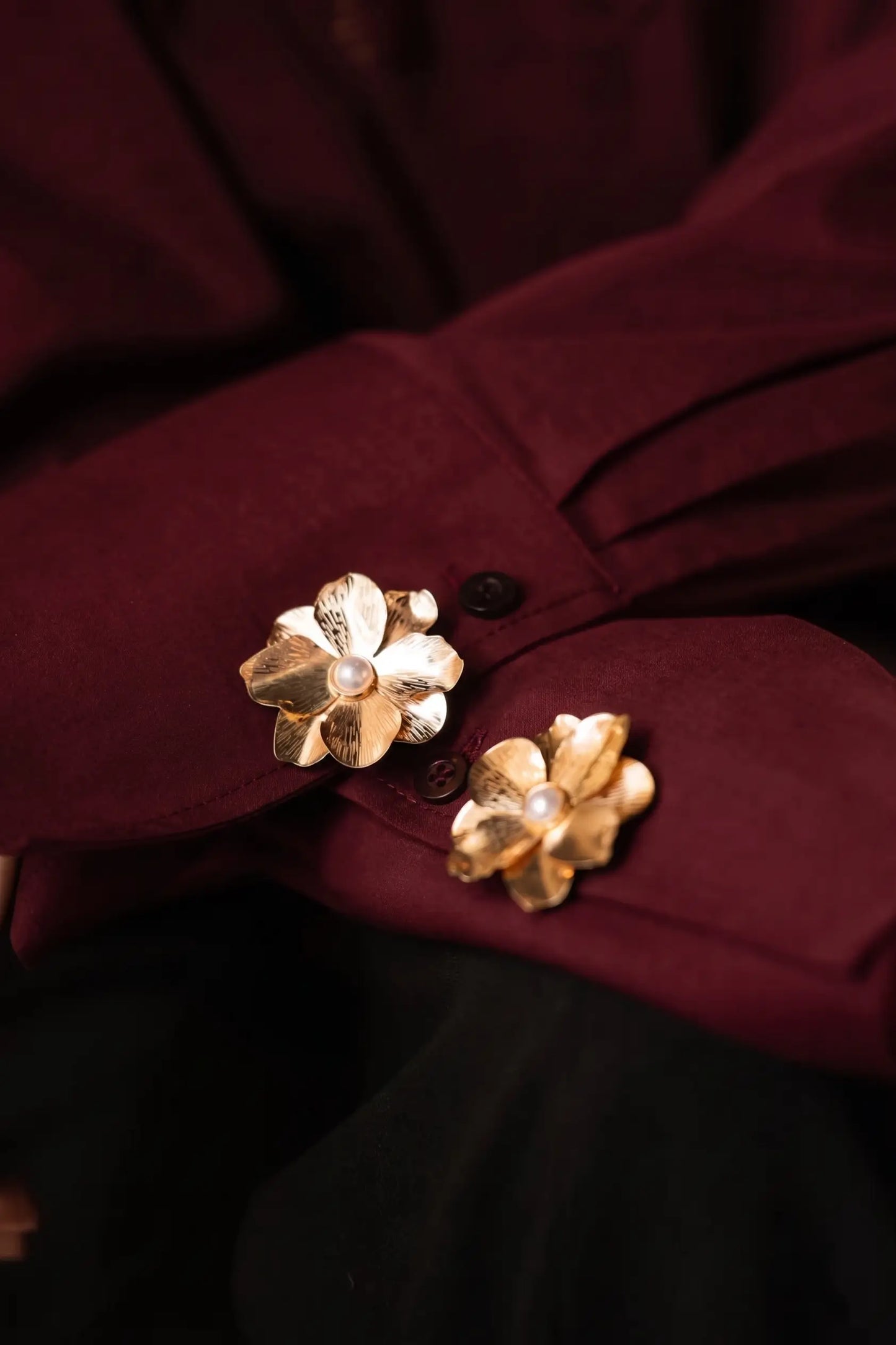 A close-up of folded burgundy shirt cuffs elegantly decorated with two textured gold floral button covers, each featuring a classic pearl center. This rich, warm-toned styling perfectly captures the timeless elegance of the old money and quiet luxury aesthetic.