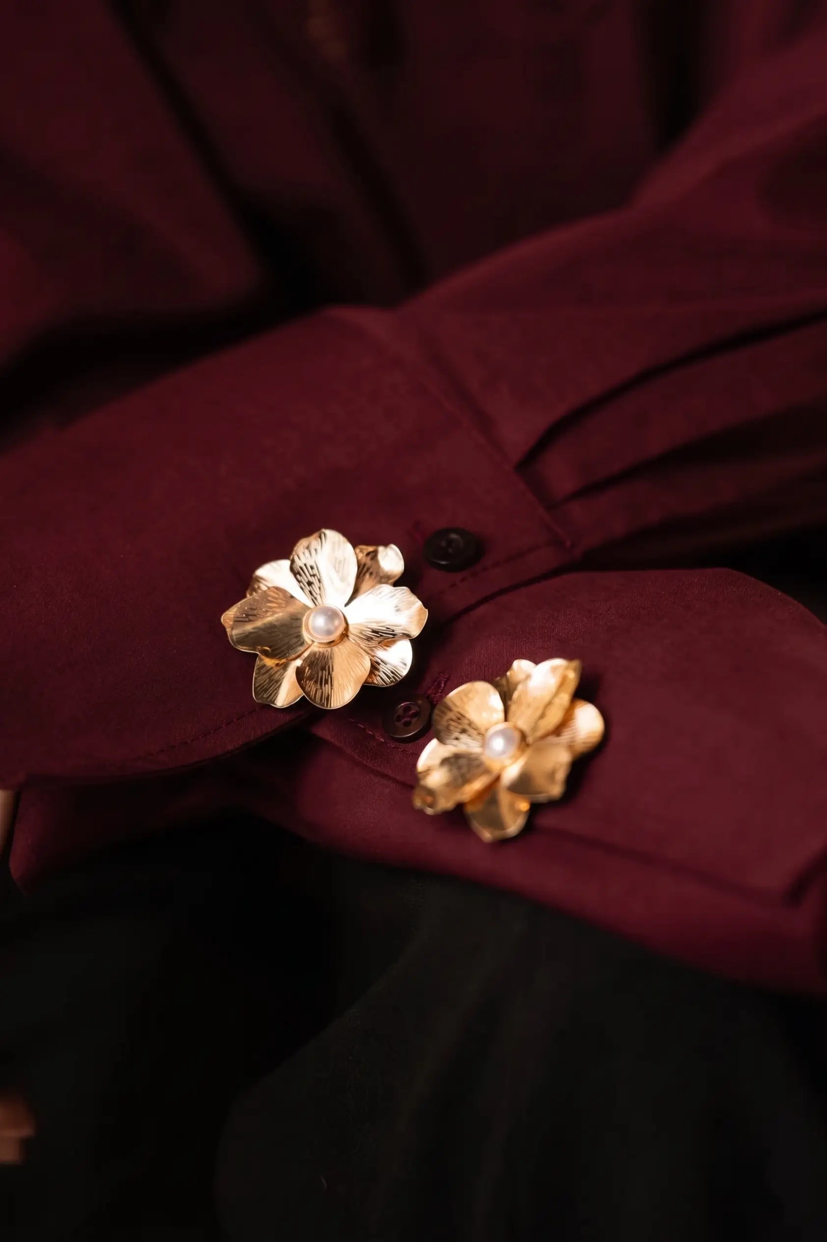 A close-up of folded burgundy shirt cuffs elegantly decorated with two textured gold floral button covers, each featuring a classic pearl center. This rich, warm-toned styling perfectly captures the timeless elegance of the old money and quiet luxury aesthetic.