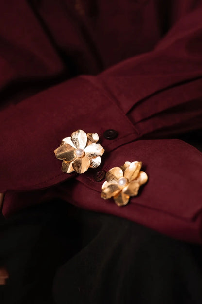 A close-up of folded burgundy shirt cuffs elegantly decorated with two textured gold floral button covers, each featuring a classic pearl center. This rich, warm-toned styling perfectly captures the timeless elegance of the old money and quiet luxury aesthetic.