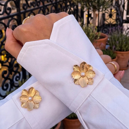 An elegant lifestyle shot of a woman's crossed arms wearing a crisp white shirt with folded cuffs. The cuffs are beautifully styled with two textured gold floral button covers featuring a pearl center. Set against a scenic city balcony at sunset, this image perfectly captures the old money aesthetic and quiet luxury fashion.