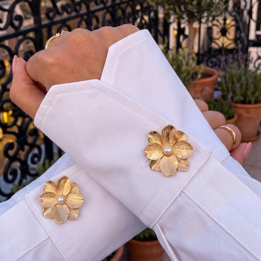 An elegant lifestyle shot of a woman's crossed arms wearing a crisp white shirt with folded cuffs. The cuffs are beautifully styled with two textured gold floral button covers featuring a pearl center. Set against a scenic city balcony at sunset, this image perfectly captures the old money aesthetic and quiet luxury fashion.