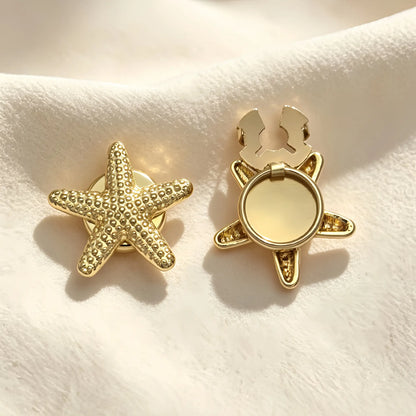 A pair of light luxury gold starfish button covers resting on soft cream fabric under warm sunlight. The left piece features a detailed, textured starfish design, while the right displays the open gold hinge mechanism, perfect for an effortless, no-sew nautical cuff upgrade.