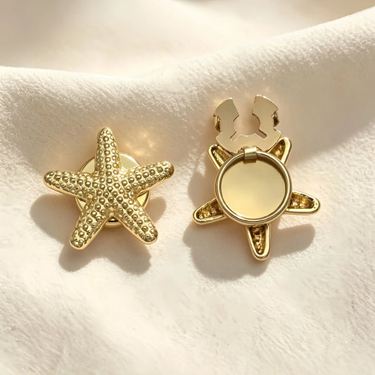 A pair of light luxury gold starfish button covers resting on soft cream fabric under warm sunlight. The left piece features a detailed, textured starfish design, while the right displays the open gold hinge mechanism, perfect for an effortless, no-sew nautical cuff upgrade.