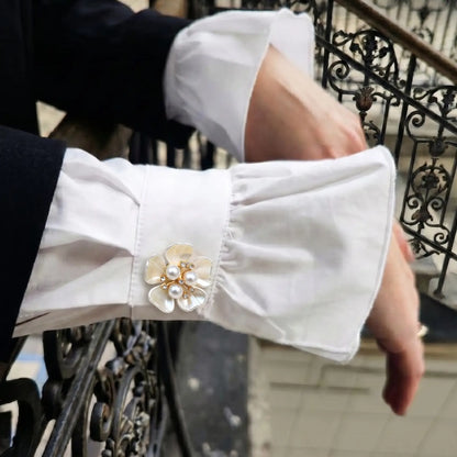 A person resting their hands on an iron balcony railing, wearing a white shirt with ruffled cuffs fastened by a white enamel floral button cover. A beautiful everyday French-style cuff upgrade for street style fashion.