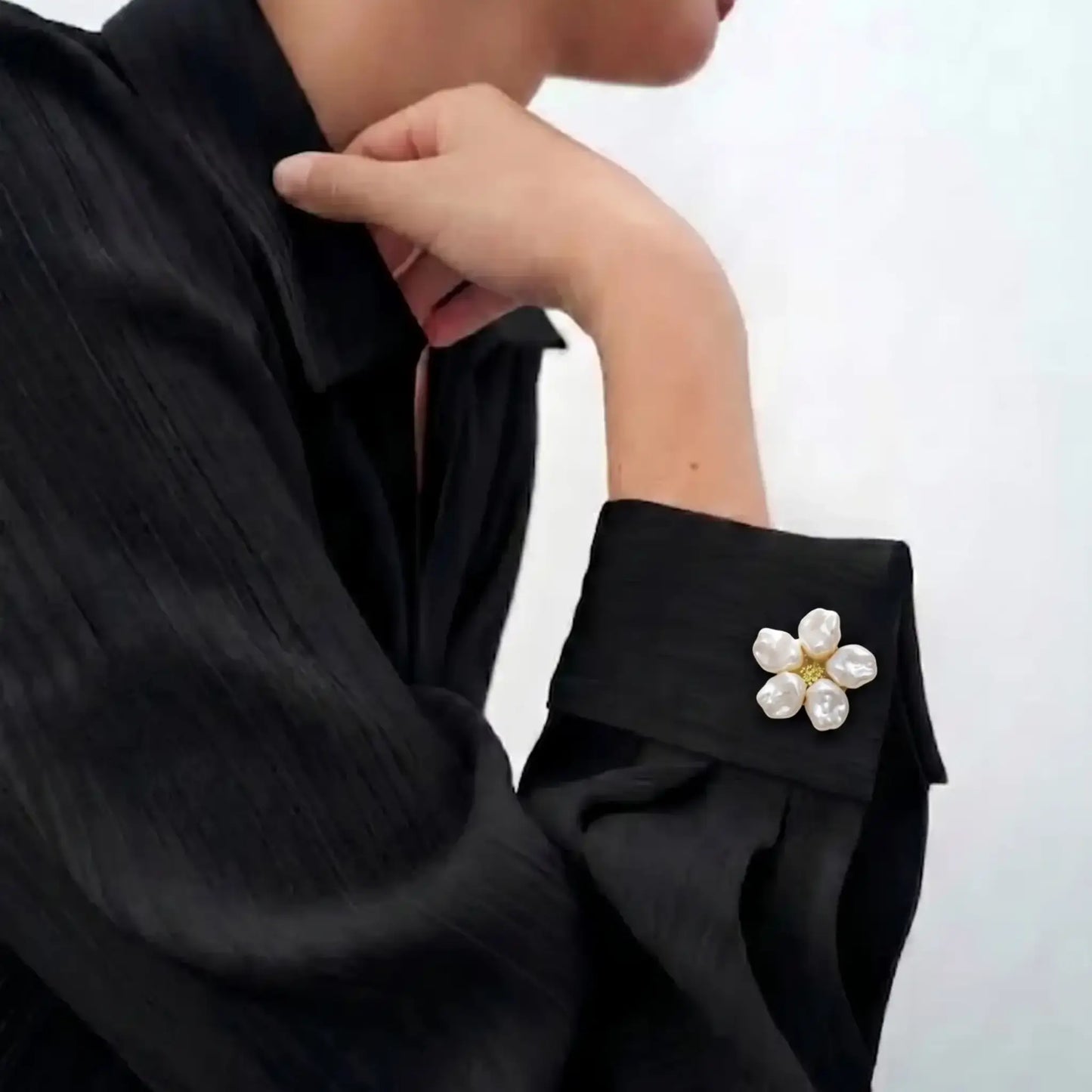 An elegant lifestyle image of a woman in a modern, dark-toned shirt, featuring a realistically small white pearl floral button cover on the cuff. This refined faux cufflink perfectly mimics the size of a standard button, seamlessly adding a touch of sophisticated, everyday fashion to the outfit.